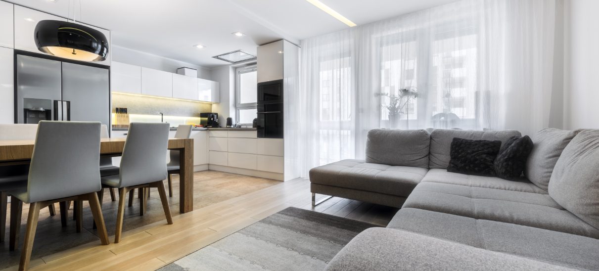 Bright apartment living area with a grey corner sofa, dining table and white kitchen