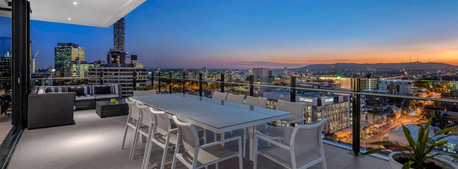Penthouse balcony with a long outdoor dining table and lounge overlooking the city at dusk