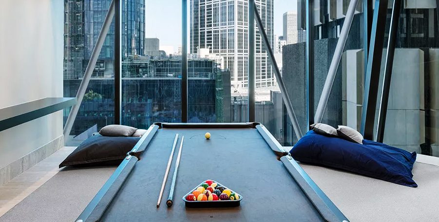Pool table with cues and racked balls beside floor-to-ceiling windows onto city towers