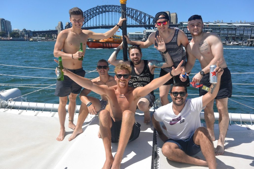 A crew of bucks on the deck of a boat with beers up, the Sydney Harbour Bridge behind them