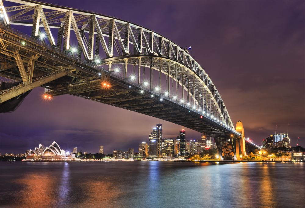 Sydney Harbour Bridge lit up at night, with the Opera House and the city lights across the water