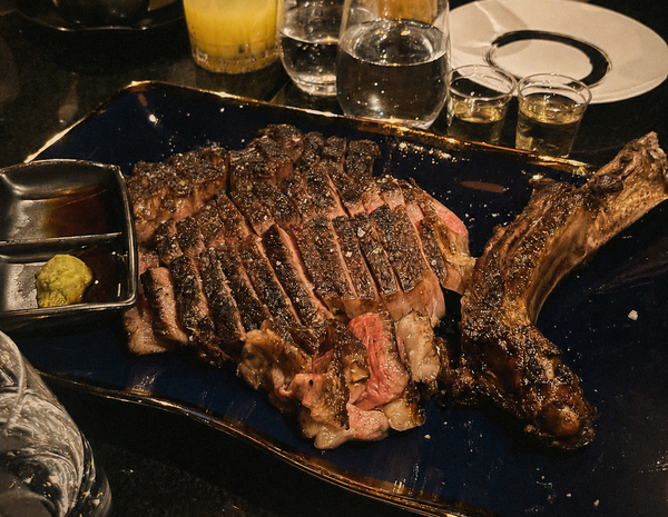 A sliced tomahawk steak on a dark plate beside drinks