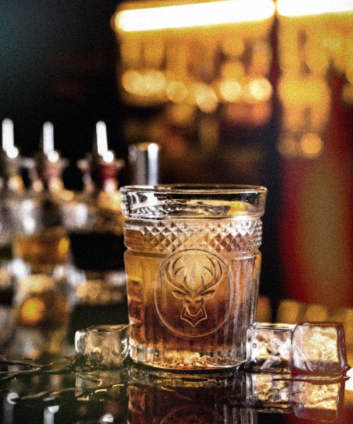 A glass of whisky on a bar with bottles glowing behind it
