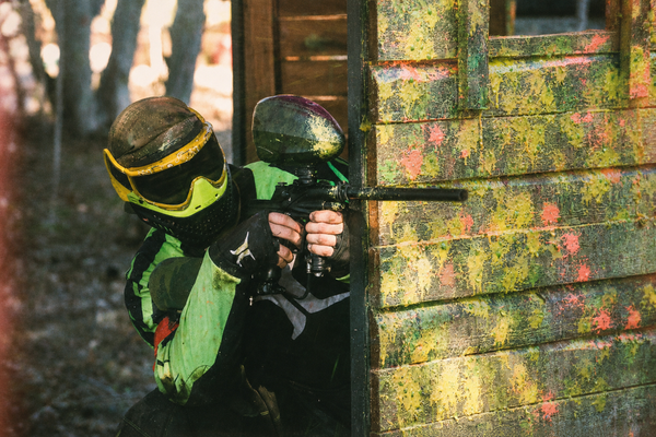 A paintballer in a green jersey aiming from behind a splattered wall