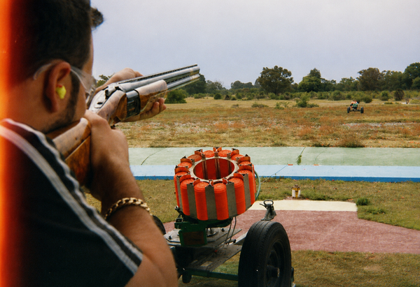 A shooter in ear protection aiming a shotgun beside a loaded clay trap